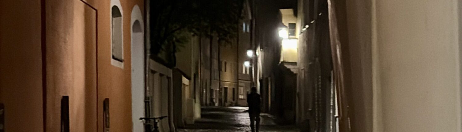 Dunkle Gasse mit historischen Gebäuden und Laternen, die den Weg beleuchten.