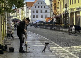 Denn wir wissen, was wir tun 5 Ein Puppenspieler mit einer Marionette auf einer gepflasterten Straße in einer Stadt, umgeben von historischen Gebäuden.