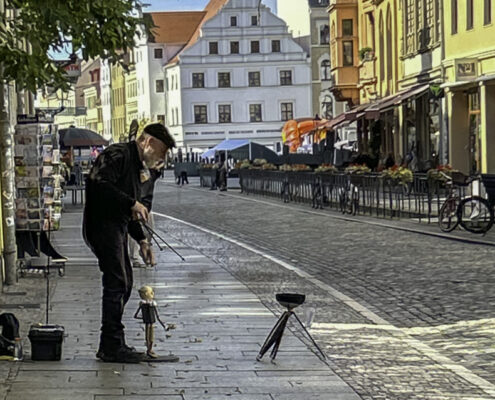 Ein Puppenspieler mit einer Marionette auf einer gepflasterten Straße in einer Stadt, umgeben von historischen Gebäuden.