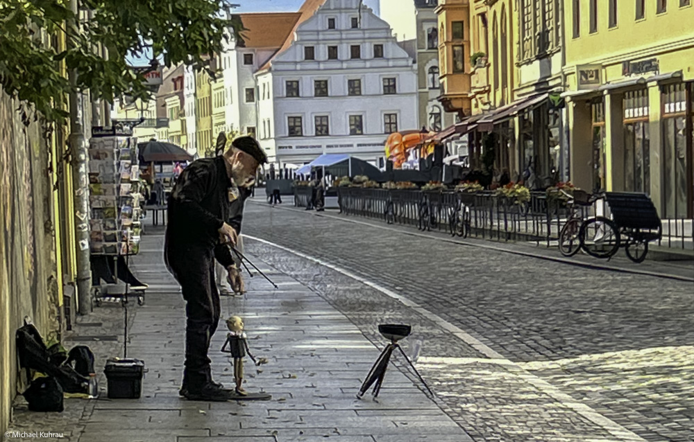 Ein Puppenspieler mit einer Marionette auf einer gepflasterten Straße in einer Stadt, umgeben von historischen Gebäuden.