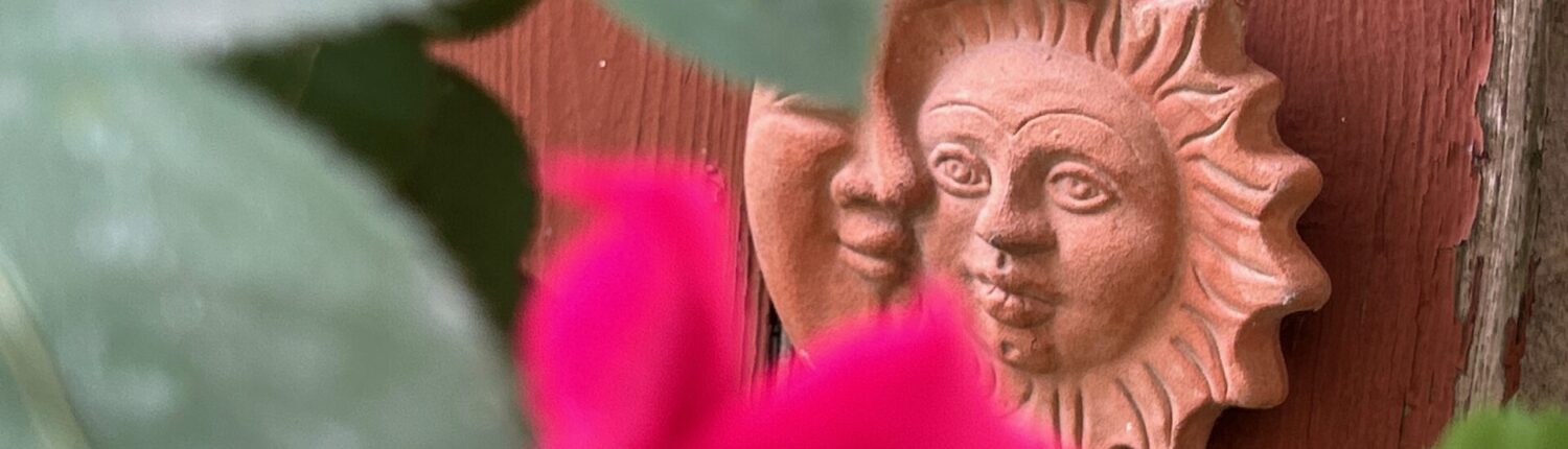 Terrakotta-Sonnensymbol mit zwei Gesichtern, das an einer Wand befestigt ist, umgeben von grünen Blättern und einer pinken Blüte.