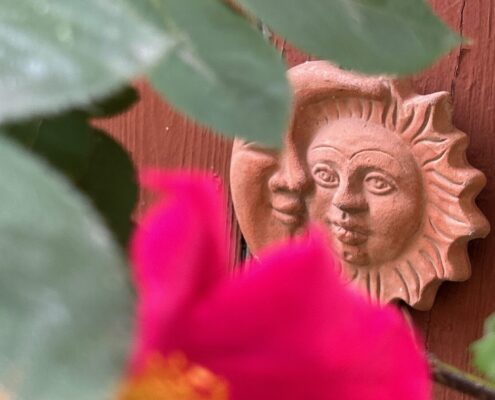 Terrakotta-Sonnensymbol mit zwei Gesichtern, das an einer Wand befestigt ist, umgeben von grünen Blättern und einer pinken Blüte.