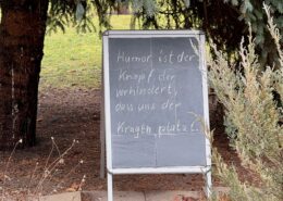 Tafel mit der Aufschrift 'Humor ist der Knopf, der verhindert, dass uns der Kragen platzt.' in einem Garten.