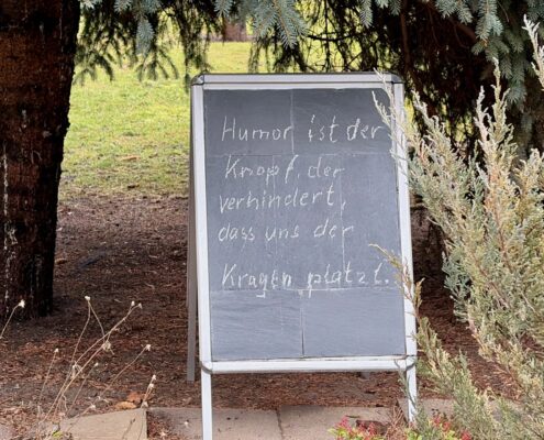 Tafel mit der Aufschrift 'Humor ist der Knopf, der verhindert, dass uns der Kragen platzt.' in einem Garten.