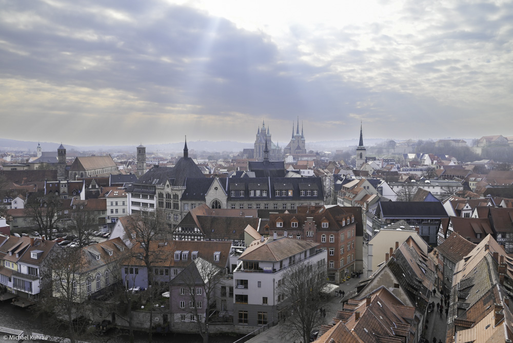Panorama der Stadt Erfurt mit dicht gedrängten roten Ziegeldächern, markanten Kirchtürmen und historischen Gebäuden unter einem Himmel mit Sonnenstrahlen