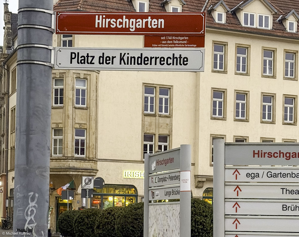 Erfahrungen und Fotos aus Erfurt 32 Erfurt Hirschgarten Straßenschilder am Platz der Kinderrechte – rotes Schild über weißem Schild vor beige Gebäuden