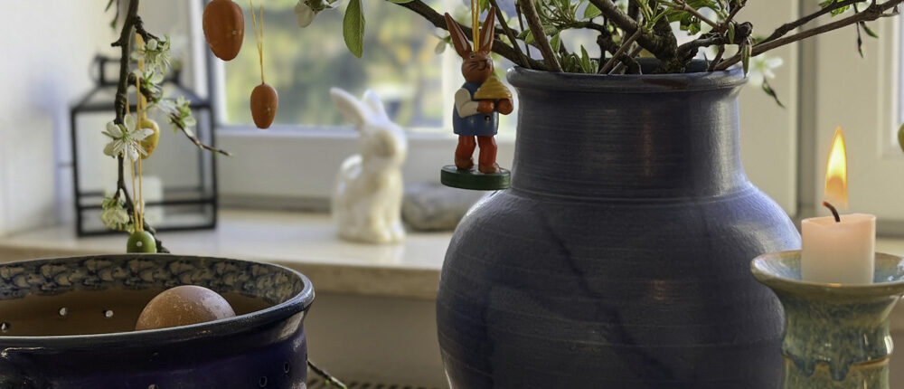 Blaues Keramikvase mit grünem Zweigeschmuck, hängende rote Ostereier, Kerzenhalter und eine kleine Bunny-Figur im Hintergrund – Kuhrau.de