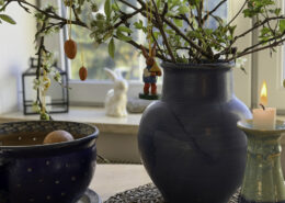 Blaues Keramikvase mit grünem Zweigeschmuck, hängende rote Ostereier, Kerzenhalter und eine kleine Bunny-Figur im Hintergrund – Kuhrau.de