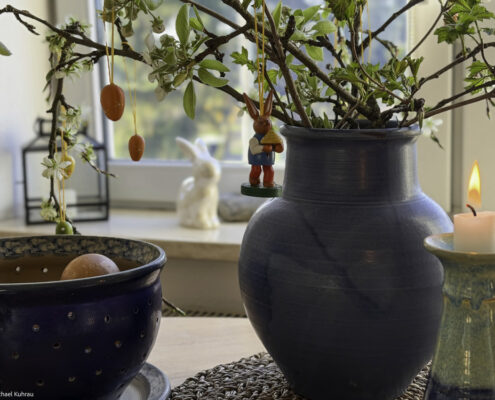 Keiner sagt Halt - Michael's Gedanken zu Ostern 2026 1 Blaues Keramikvase mit grünem Zweigeschmuck, hängende rote Ostereier, Kerzenhalter und eine kleine Bunny-Figur im Hintergrund – Kuhrau.de