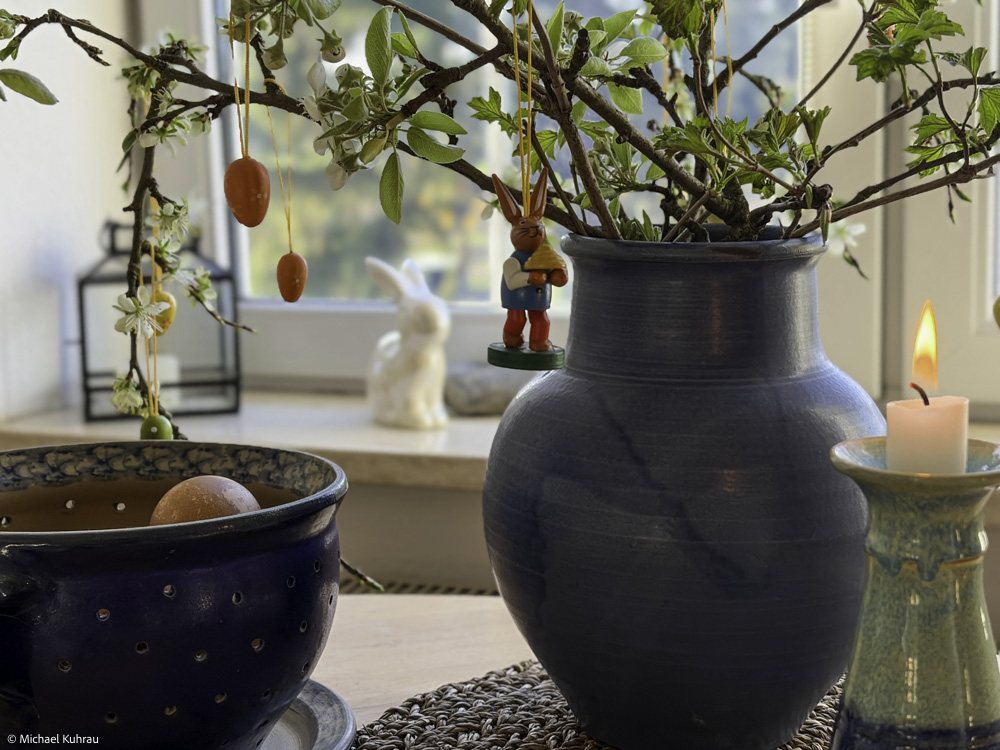 Keiner sagt Halt - Michael's Gedanken zu Ostern 2026 2 Blaues Keramikvase mit grünem Zweigeschmuck, hängende rote Ostereier, Kerzenhalter und eine kleine Bunny-Figur im Hintergrund – Kuhrau.de