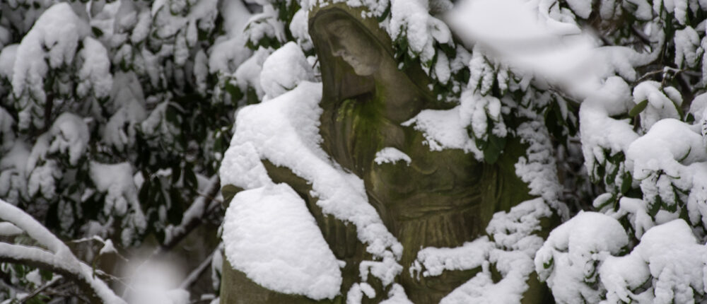 Traurig sein - Eine verwitterte Skulptur mit Kapuze steht zwischen schneebedeckten Ästen und trägt dicke Schneeschichten.
