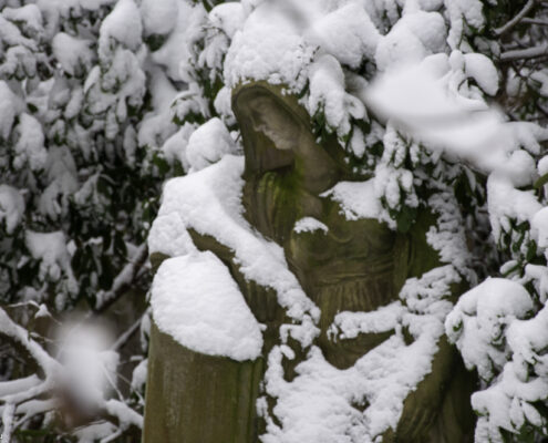 Traurig sein 1 Traurig sein - Eine verwitterte Skulptur mit Kapuze steht zwischen schneebedeckten Ästen und trägt dicke Schneeschichten.