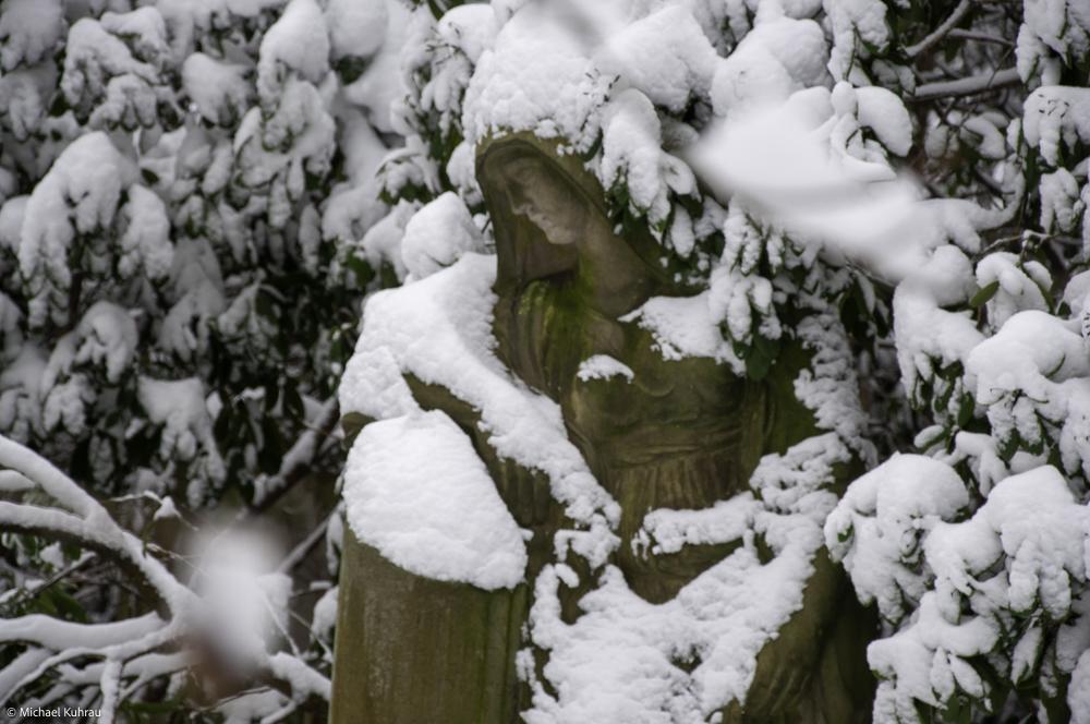 Traurig sein 1 Traurig sein - Eine verwitterte Skulptur mit Kapuze steht zwischen schneebedeckten Ästen und trägt dicke Schneeschichten.